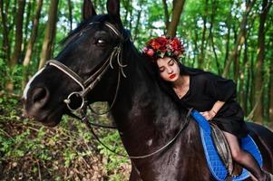 Mystical girl in wreath wear in black at horse in wood. photo