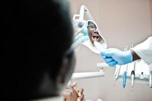 African american man patient in dental chair. Dentist office and doctor practice concept. Professional dentist helping his patient at dentistry medical and showing to him a mirror. photo