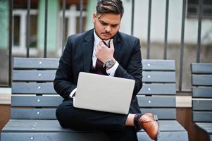 Stylish indian businessman in formal wear sitting on bench with laptop. photo