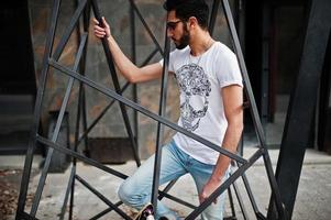 Street style arab man in eyeglasses with longboard posed inside metal pyramid construction. photo