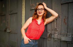 Attractive redhaired woman in eyeglasses, wear on red blouse and jeans skirt posing. photo