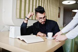 Arab man wear on black jeans jacket and eyeglasses sitting in cafe, read book and drink coffee. Stylish and fashionable arabian model guy. Hands of waiter. photo