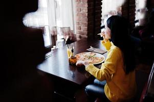 chica morena divertida en suéter amarillo comiendo pizza en el restaurante. foto