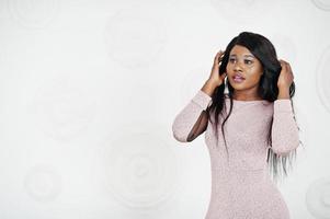 Fashionable african american model woman in pink brilliant evening dress posed against white decorative wall. photo