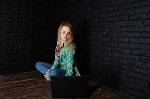 Stylish blonde girl in jacket and jeans with laptop against brick black wall at studio. photo