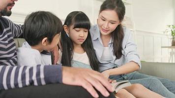 famiglia tailandese asiatica, papà adulto, mamma e bambini felicità vita domestica attività rilassanti e lettura di libri insieme, tempo libero sul divano in una stanza bianca, fine settimana incantevole, benessere stile di vita domestico. video