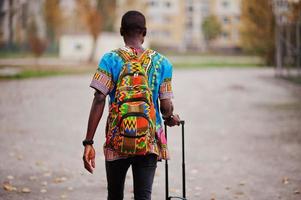 African man in africa traditional shirt on autumn park with backpack and suitcase. Emigrant traveler. photo