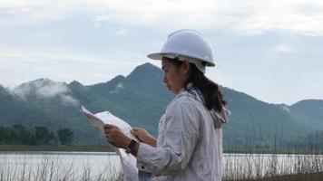 donna ecologista di ingegneria in un casco che tiene un progetto si trova sulla riva di un fiume per sviluppare una diga idroelettrica per generare elettricità. concetti di energia pulita e tecnologia. video