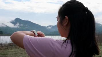 una mujer asiática corre por el campo junto al río por la mañana y comprueba su pulso en su reloj mientras descansa. concepto de estilo de vida saludable deportivo y recreativo. video