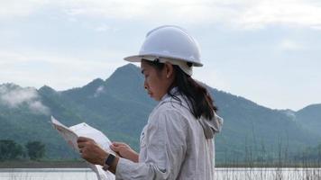 Engineering ecologist woman in a helmet holding a map stands on the bank of a river to develop a hydroelectric dam to generate electricity. Clean energy and Technology concepts. video