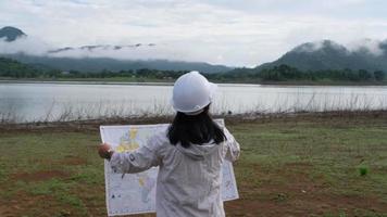 Engineering ecologist woman in a helmet holding a map stands on the bank of a river to develop a hydroelectric dam to generate electricity. Clean energy and Technology concepts. video