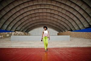 Amazing african american model woman in green pants and black hat posed outdoor against arena hall. photo