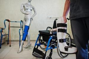 Prosthetist man with prosthetic leg in laboratory against wheelchair. photo