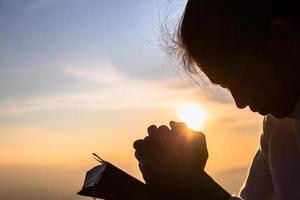 Silhouette of christian young woman praying with a  cross and open the bible at sunrise, Christian Religion concept background. photo