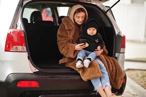 Young mother and child sitting in the trunk of a car and looking at mobile phone. Safety driving concept. photo