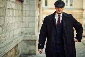 retrato de un hombre de negocios árabe inglés retro de los años 20 con abrigo oscuro, traje, corbata y gorra plana. foto