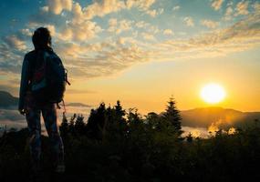 Young fit happy woman stand on viewpoint after reach top in mountains. Carefree tourist woman looking at sun enjoying landscape. Girl traveler on top of mountain in rays of sunset photo