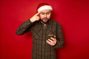 thoughtful serious man in xmas hat reads on phone holds index finger to eyebrow tries to understand conditions isolated studio background concept - holidays, technology, social networks, communication photo