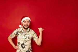 a portrait of a joyful smiling man in a Christmas hat and a tourist shirt points with his thumb to side at empty space for text. isolated on red studio background. concept - travel, new year, people photo