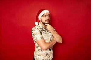 viajero turista serio y reflexivo con sombrero de navidad, poner la mano en la barbilla en pose de reflexión a dónde ir a volar para descansar, celebrar el concepto de fondo rojo de estudio aislado de año nuevo - toma de decisiones foto