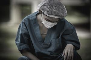 Tired depressed female asian scrub nurse wears face mask blue uniform sits on hospital floor,Young woman doctor stressed from hard work photo