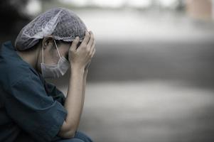 Tired depressed female asian scrub nurse wears face mask blue uniform sits on hospital floor,Young woman doctor stressed from hard work photo