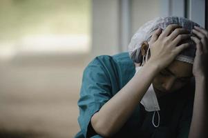 Tired depressed female asian scrub nurse wears face mask blue uniform sits on hospital floor,Young woman doctor stressed from hard work photo