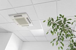 Low angle of assette Air Conditioner on ceiling in modern light office or apartment with green ficus plant leaves. Indoor air quality photo