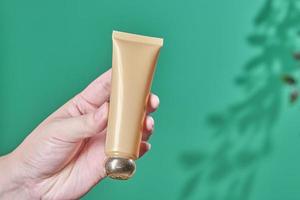 Female hand holds cosmetic product in tube, glass bottle, lotion, anti-aging moisturizer, facial essential oil or serum on green background with light shadow nature. Beauty cosmetic skincare concept. photo