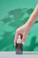 Female hand holds cosmetic product in tube, glass bottle, lotion, anti-aging moisturizer, facial essential oil or serum on green background with light shadow nature. Beauty cosmetic skincare concept. photo