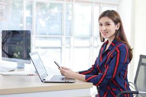Asian woman holds the mobile phone on her hands with smiling face in the office while there are laptop and computer put on the table. photo