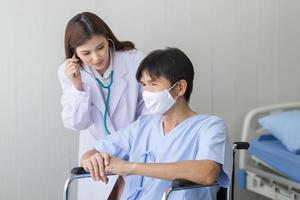 Asian woman doctor use a stethoscope to check the lung rhythm of a male patient who wear face mask while he sit on a wheelchair at hospital. photo