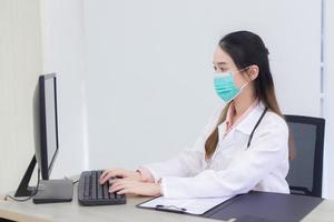 Asian woman doctor is typing on keyboard to record information into computer while she wears medical face mask in hospital. photo