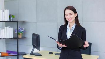 Professional Asian business woman in black suit confident smiles happily while she works and holds clipboard in office. photo