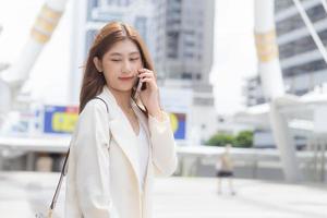 una joven mujer de negocios asiática con traje blanco sonríe y sostiene un teléfono inteligente en sus manos en un feliz día de trabajo en la ciudad como fondo. foto