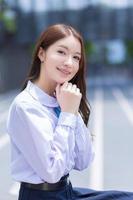 Beautiful Asian high school student girl in the school uniform with braces on her teeth sits and smiles confidently while she looks at the camera happily with the building in the background. photo