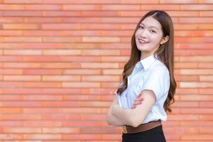 retrato de un estudiante tailandés adulto con uniforme de estudiante universitario. hermosa chica asiática de pie con los brazos cruzados sobre un fondo de ladrillo. foto