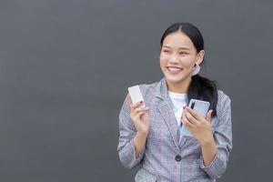 Professional Asian woman in a Brown striped blazer smiles happily successful while holds smartphone and credit card among on the dark gray wall.. photo
