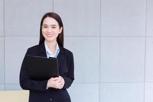 una joven trabajadora profesional asiática con traje negro sostiene un portapapeles en sus manos y sonríe confiada en la oficina. foto