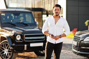 Solid asian man in white shirt and sunglasses posed near black mafia suv cars. photo