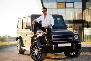 Solid asian man in white shirt and sunglasses posed near black mafia suv car. photo