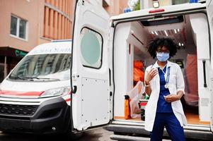 African american female paramedic in face protective medical mask standing in front of ambulance car. photo
