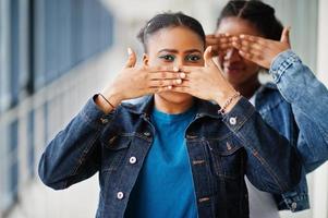 Two african woman friends in jeans jacket covered mouth and eyes with hands indoor together. photo