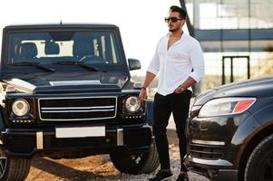 Solid asian man in white shirt and sunglasses posed near black mafia suv cars. photo