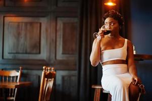 African american woman, retro hairstyle in white dress at restaurant with glass of wine. photo