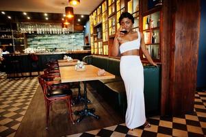 African american woman, retro hairstyle in white dress at restaurant with glass of wine. photo