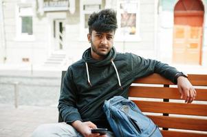 Portrait of young stylish indian man model pose in street, sitting on bench and hold smartphone at hand. photo