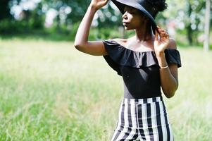 Portrait of gorgeous african american woman 20s in wear in black and white stripe pants and summer hat posing at green grass in park. photo