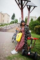 hermosa mujer afroamericana cargando segway o hoverboard con paneles solares. chica negra con símbolo de reciclaje de bolsas ecológicas de tela amarilla. foto