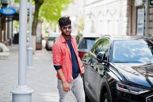 Portrait of young stylish indian man model pose in street in sunglasses against car. photo